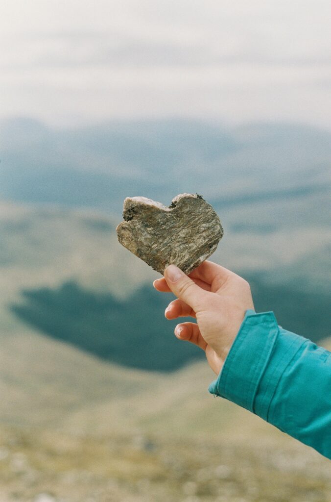 A hand holds a heart-shaped rock against a scenic Scottish Highlands backdrop.